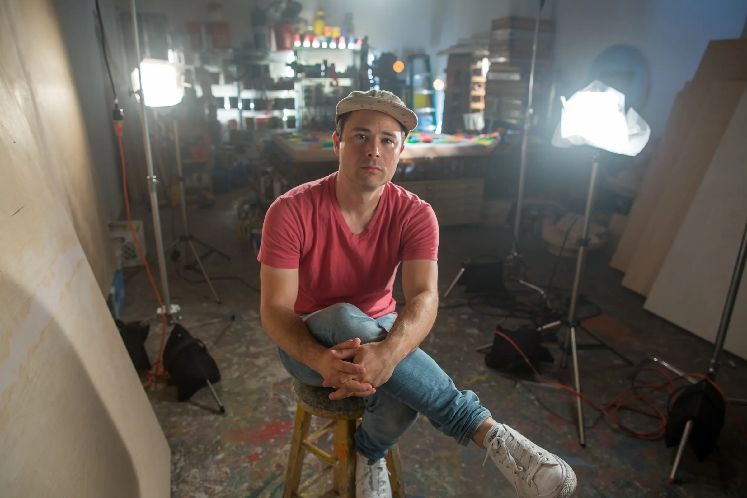 Man in a pink t-shirt seated on a stool in an art studio